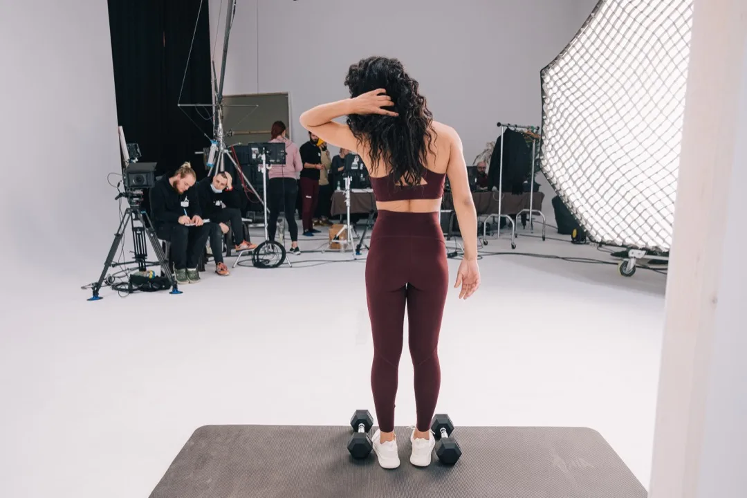 Model on set with dumbbells while the crew prepares the frame